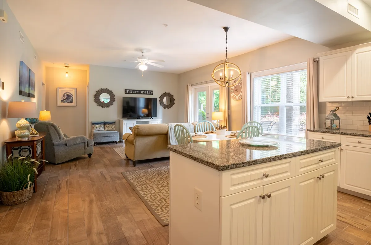 The image showcases an inviting open-concept living area with a modern kitchen island, a cozy sofa facing a large television, and a stylish dining area set against bright windows. The space feels warm, comfortable, and perfect for gathering, with plenty of natural light enhancing the relaxed, welcoming atmosphere.