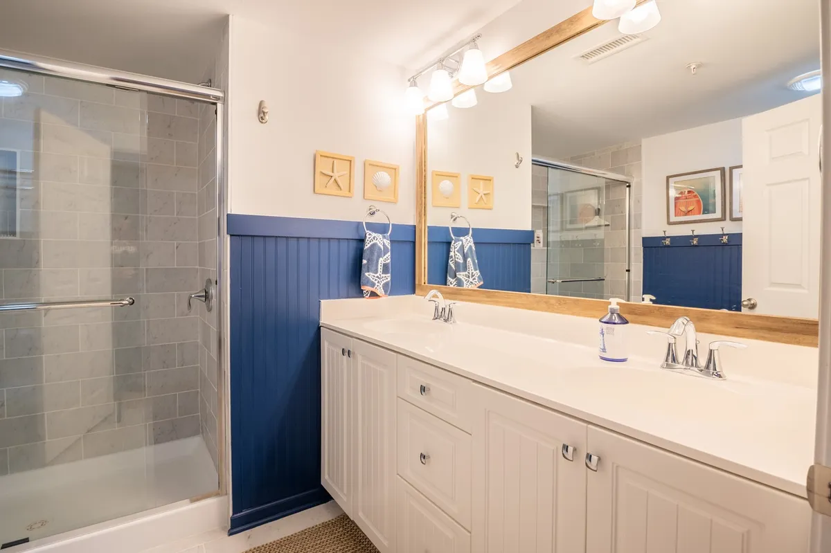 A modern bathroom featuring a double sink vanity with a large framed mirror, a decorative starfish accent, blue wainscoting, and a walk-in shower. The space is fresh and serene, ideal for relaxation after a day at the beach.