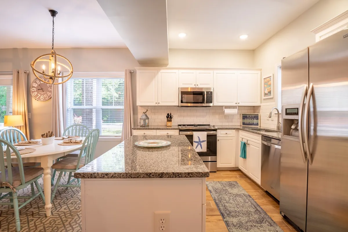 This image showcases a bright, open kitchen and dining area with a granite countertop, stainless steel appliances, and a dining table with blue chairs. The space is well-lit, inviting, and designed for both cooking and gathering. Decorative lighting and tasteful finishes add to the modern, welcoming feel.