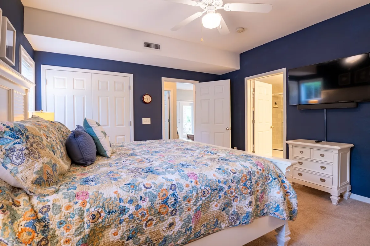 A serene bedroom with a colorful quilt on the bed, a wall-mounted TV for entertainment, and a bright, airy feel thanks to natural light flooding in. The white dresser adds ample storage for guests staying longer.