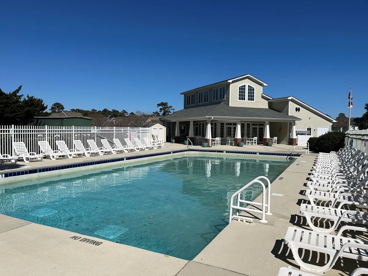 This image captures the inviting outdoor swimming pool area, framed by poolside lounge chairs and a clubhouse in the background. The scene suggests spaces for socializing, sunbathing, and enjoying recreational time in the sun, with hints of activity and relaxation.