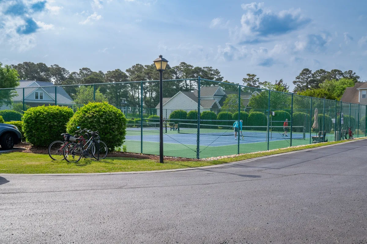 A view of well-maintained tennis courts flanked by bicycles and landscaped bushes, with a lamp post nearby. The setting feels active and communal, showcasing opportunities for group activities like tennis matches, bike rides, and neighborhood strolls.